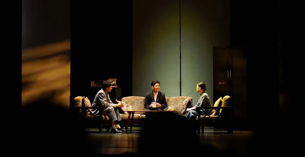 A wide view of the Finding Li Bai stage, with three actors seated around a sofa and coffee table under warm lighting