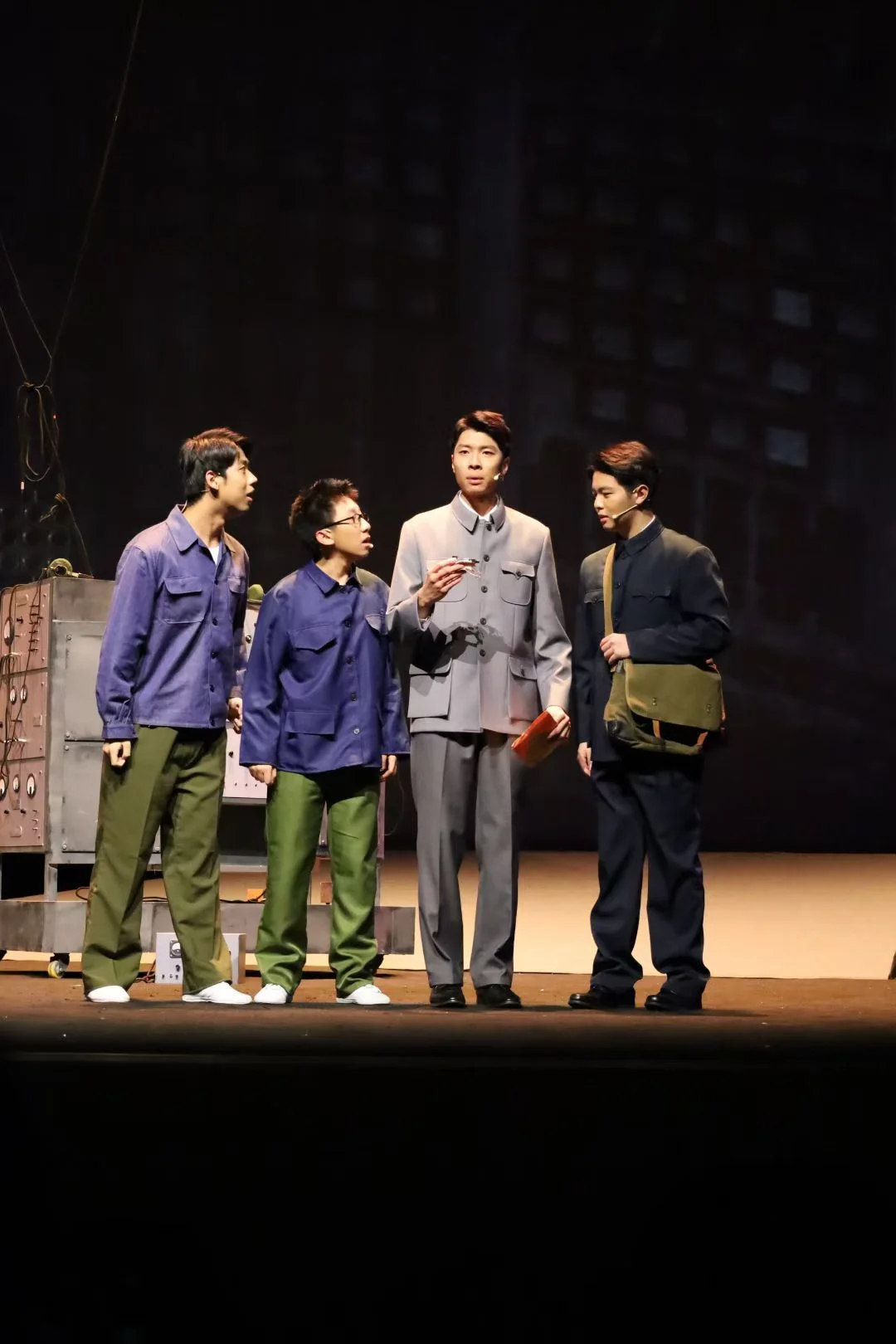 Four actors gathered on stage, with the central performer holding a pen and a red notebook