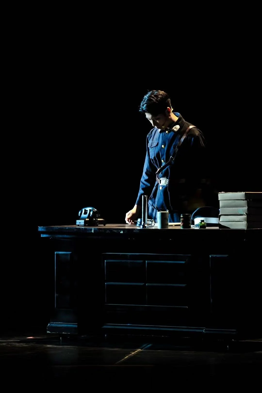 An actor in a dark uniform looking down at a desk with a telephone, cups and stacked document boxes