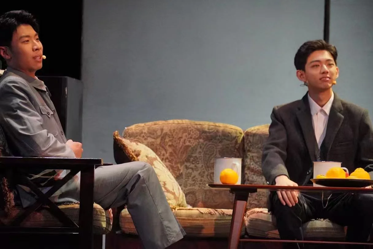 Two actors seated beside a patterned sofa, with mugs and oranges on the coffee table