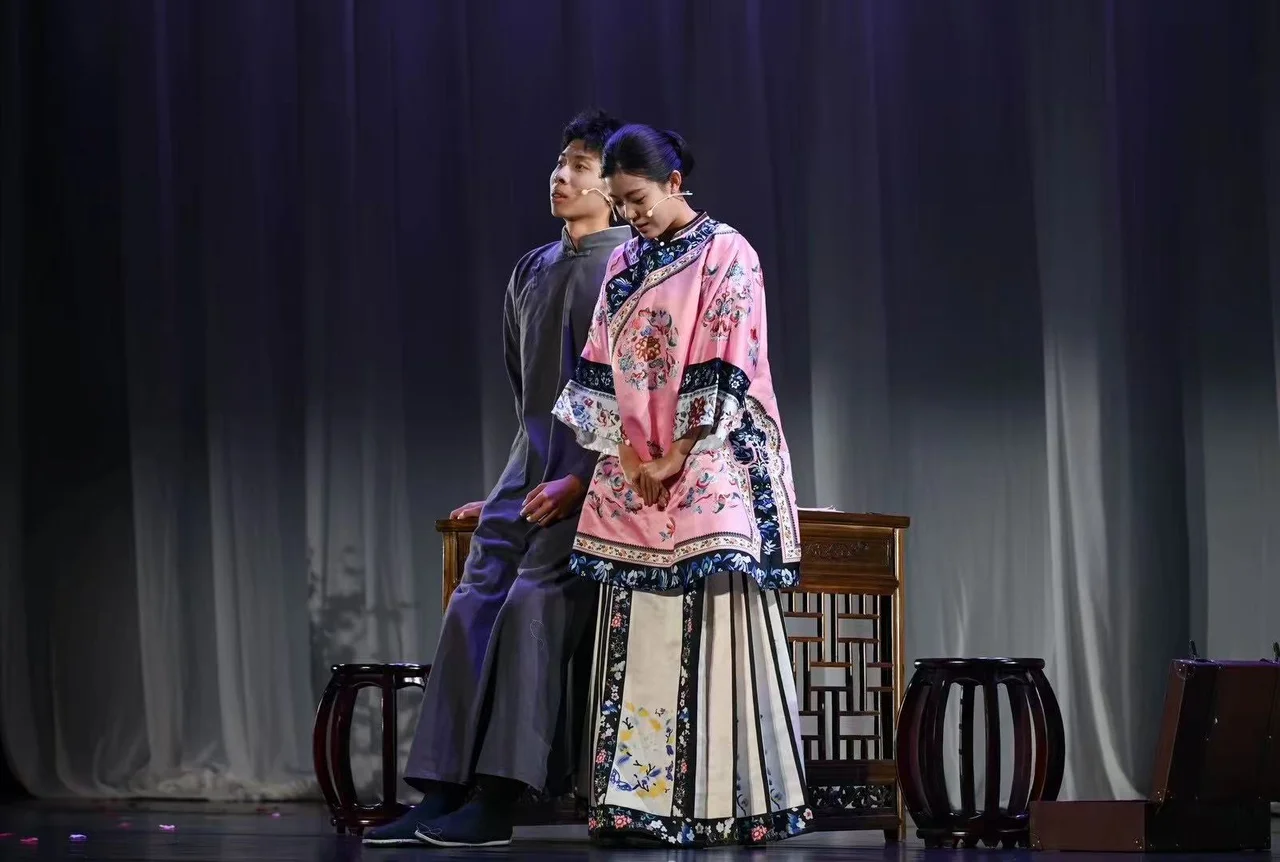 An actor in a grey changshan seated beside a wooden table, with an actor in pink embroidered clothing leaning towards him