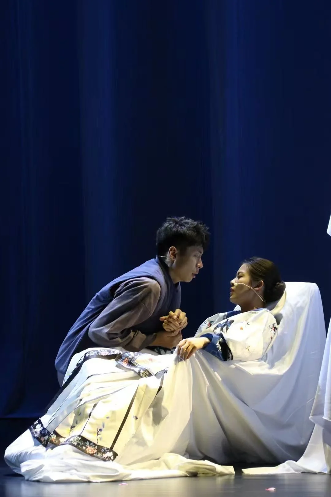 An actor in a grey changshan leaning over to hold the hand of an actor in pale embroidered clothing reclining on a white bed