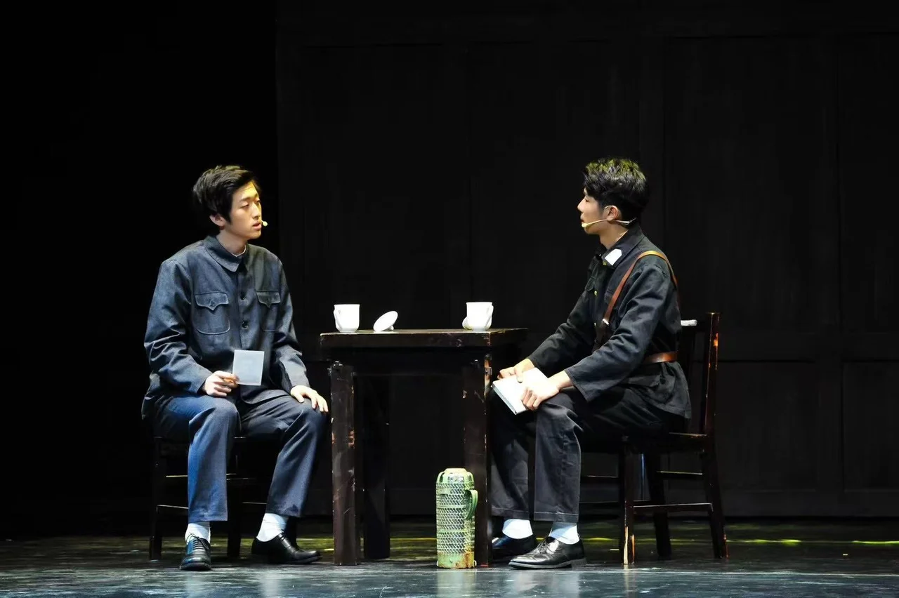 Two actors in uniform seated across a table, holding papers, with white cups between them