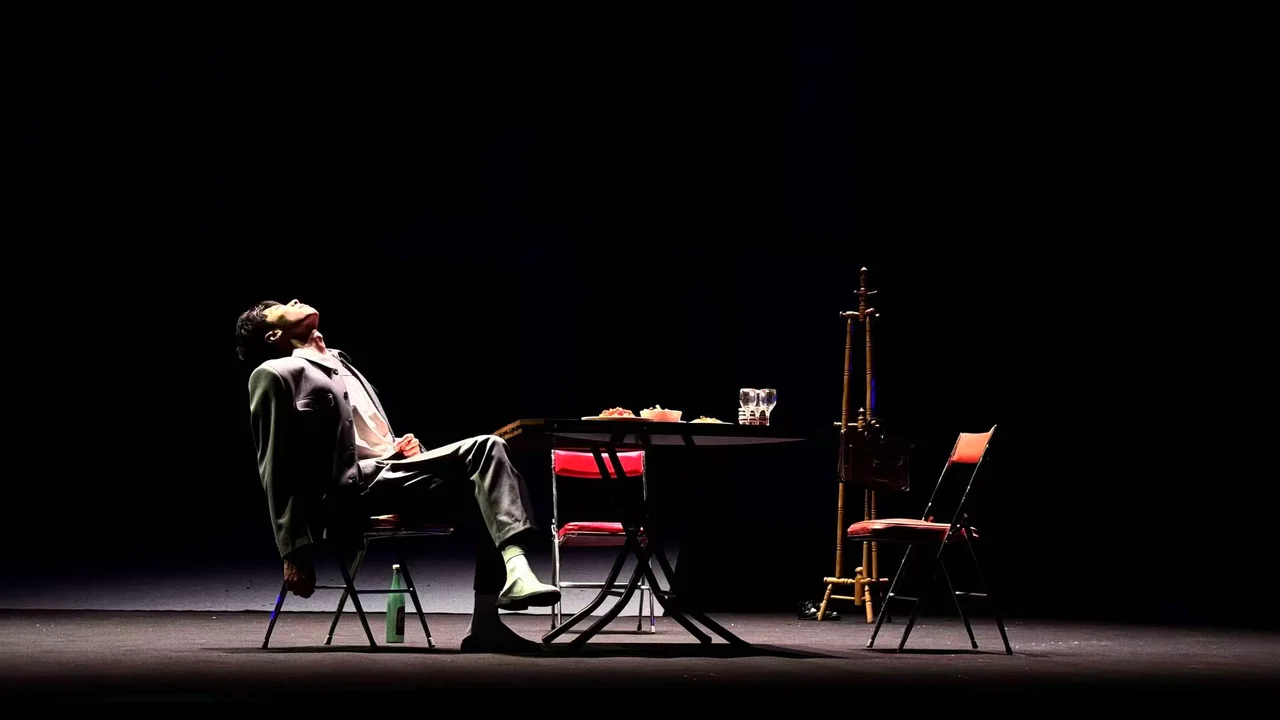 An actor leaning back at a table under stage lighting, with a coat rack and an empty chair nearby