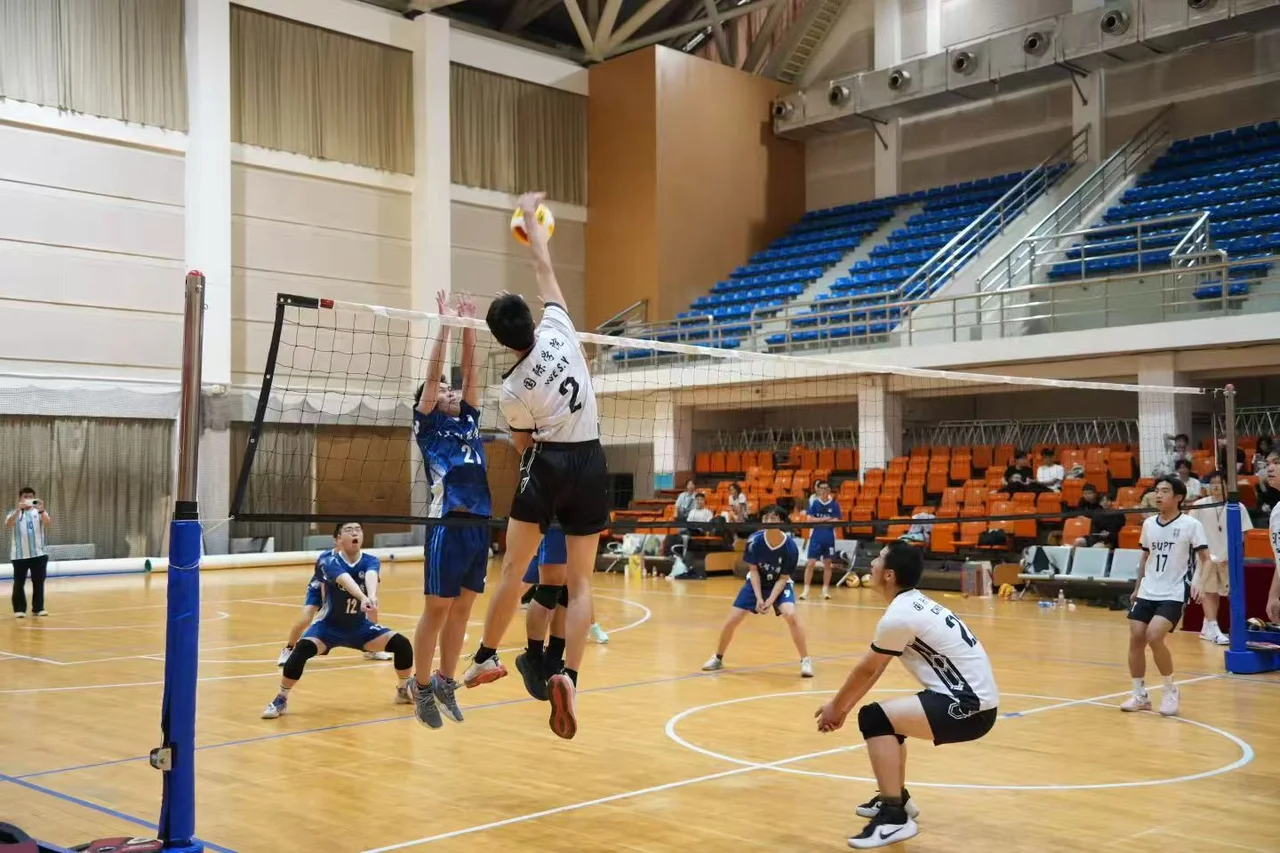 A player in a white number 2 jersey jumping to spike against a blue-shirted blocker, with other players ready for the next touch