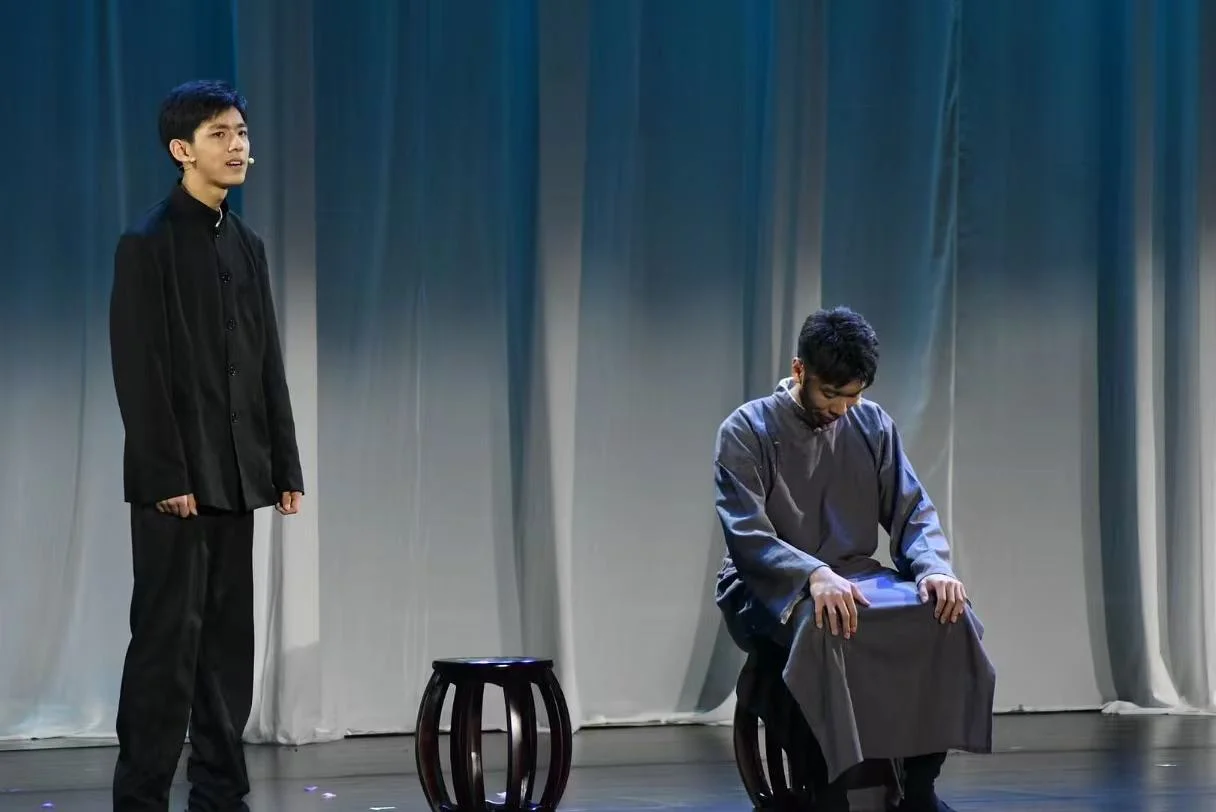An actor dressed in black standing on the left, while an actor in a grey changshan sits with his head lowered on a round stool to the right