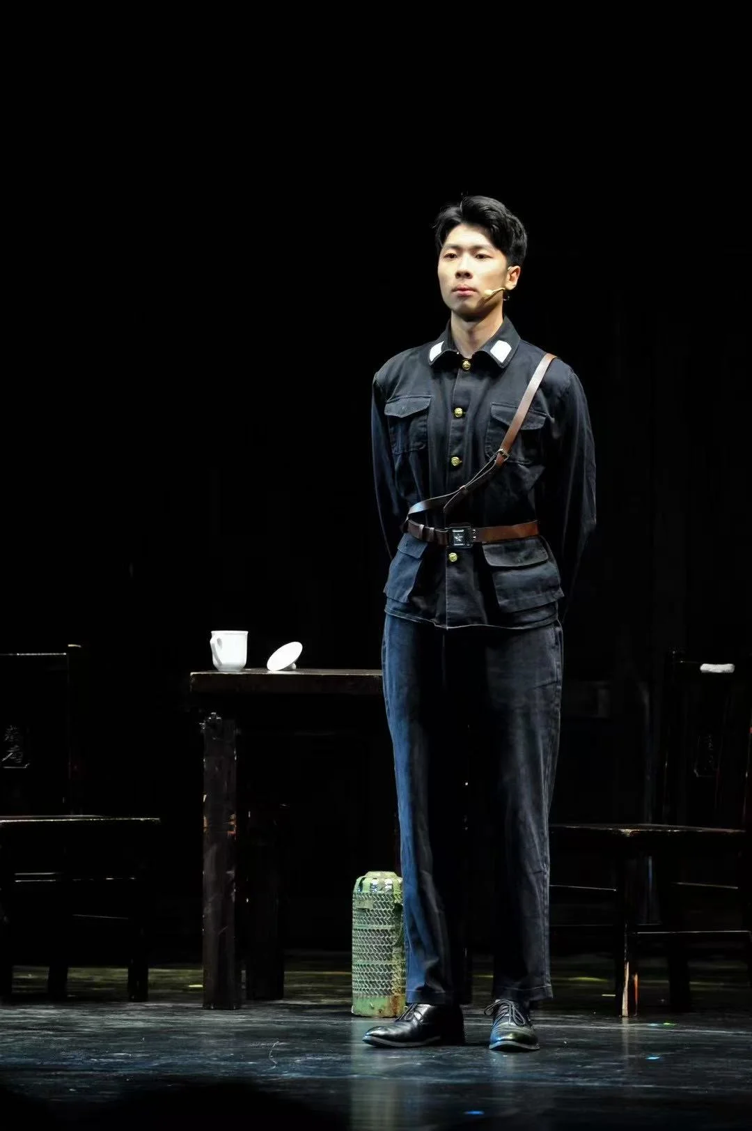 An actor in a dark uniform with a belt and shoulder strap standing on stage, with a white cup on the nearby table