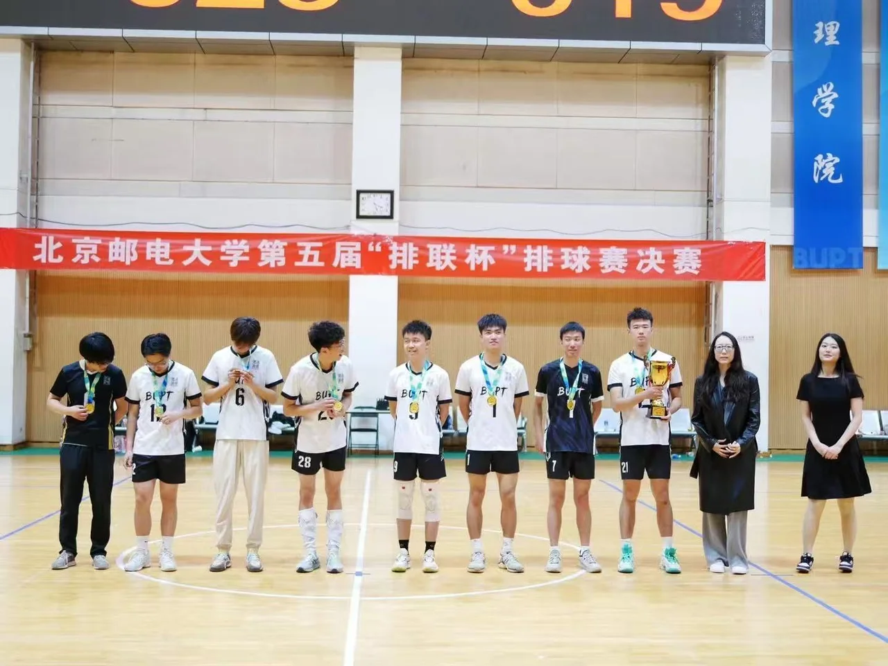 College teammates wearing medals and standing in a row beneath a campus volleyball finals banner, with one person holding a trophy