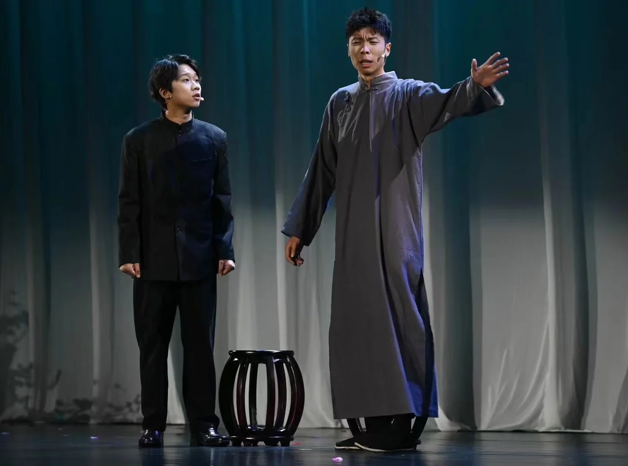 An actor in a grey changshan speaking with his arm extended, beside an actor dressed in black