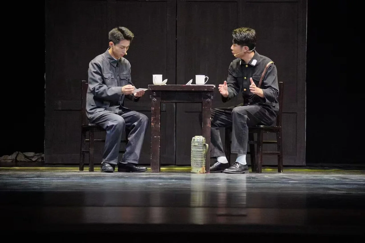 Two actors in uniform seated on opposite sides of a table, one reading a paper while the other speaks with open hands