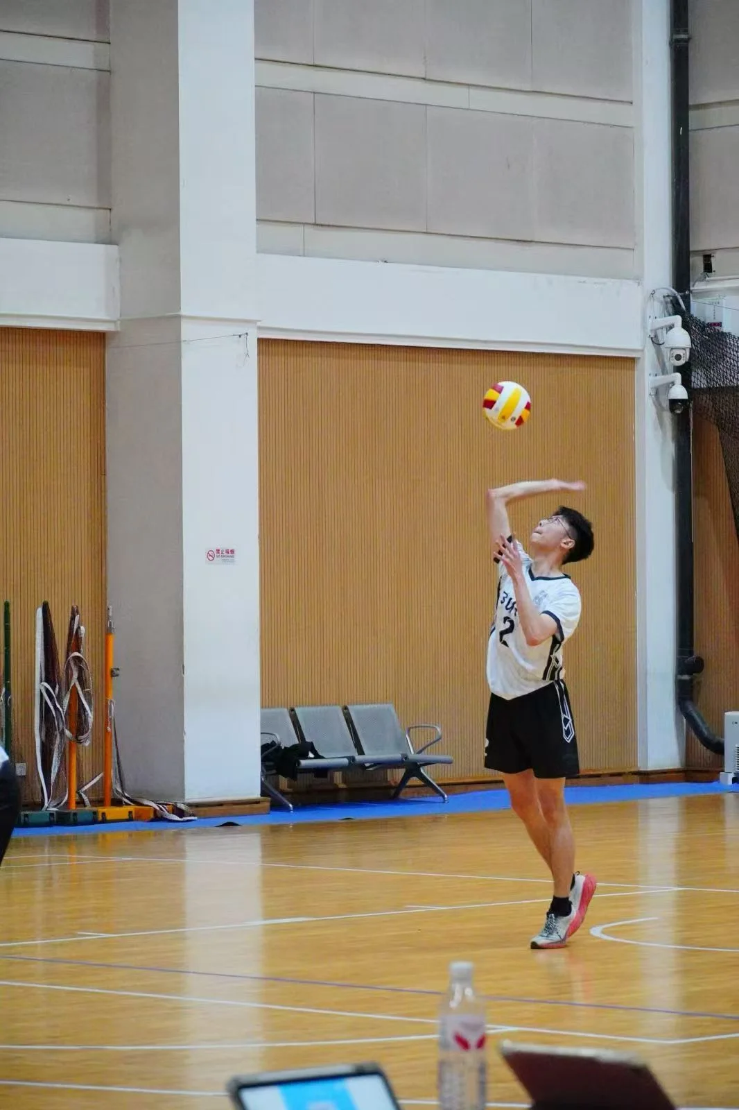 A player in a white jersey, black shorts and glasses tossing the ball to serve on an indoor volleyball court