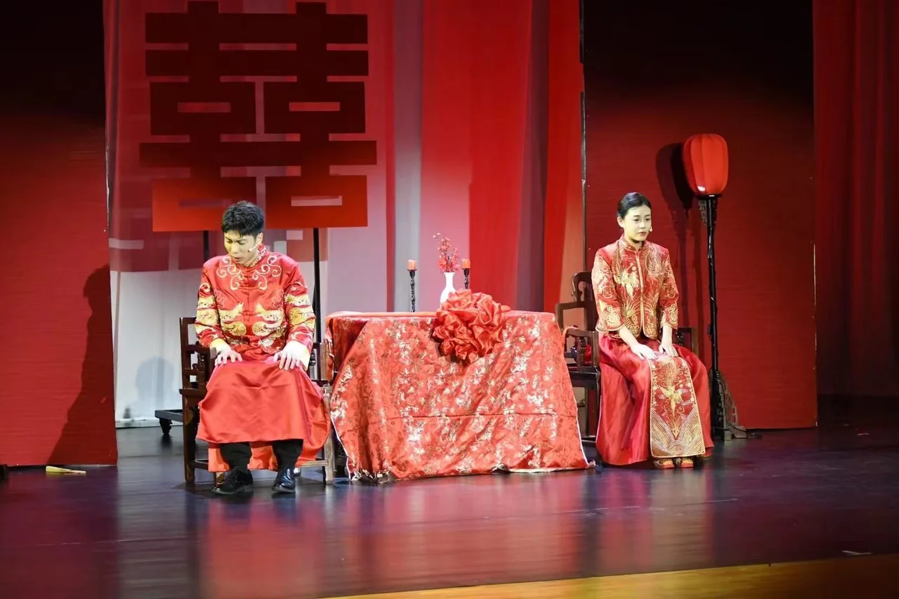 Two actors in red wedding costumes seated on opposite sides of a table, with a red double-happiness character and lantern behind them