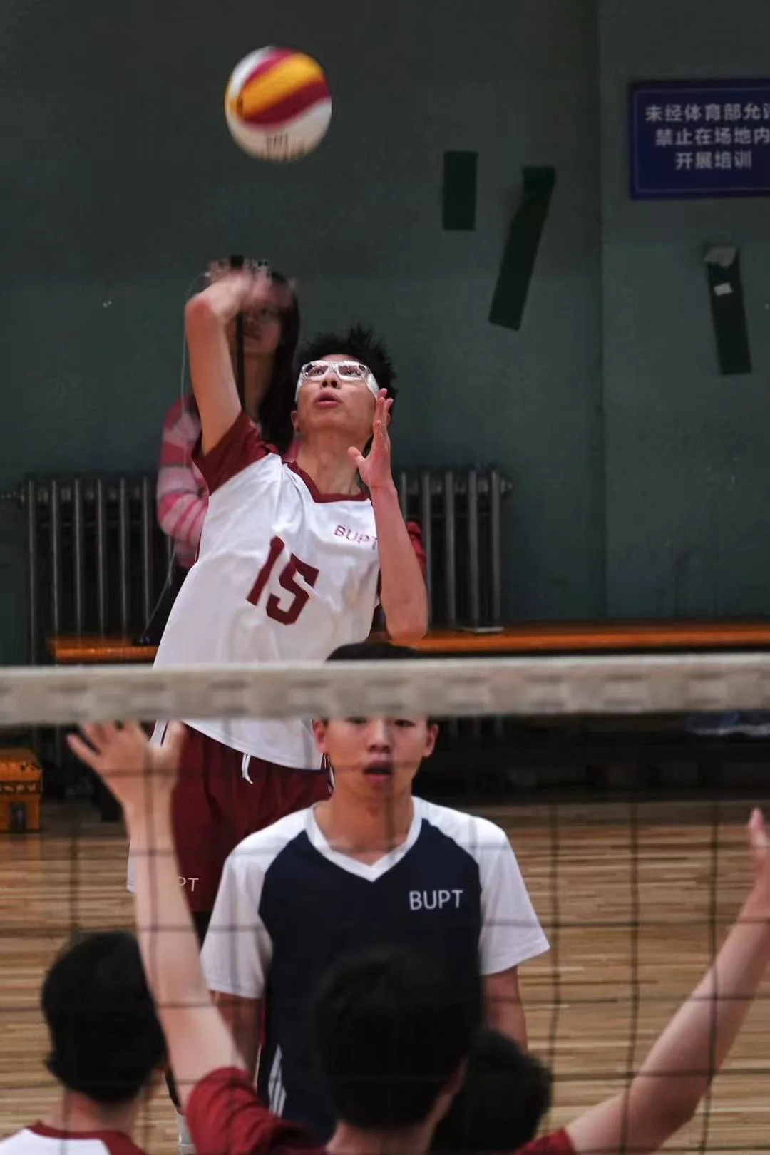 A player in a red-and-white number 15 jersey reaching up to strike the ball, with the net and other players in front