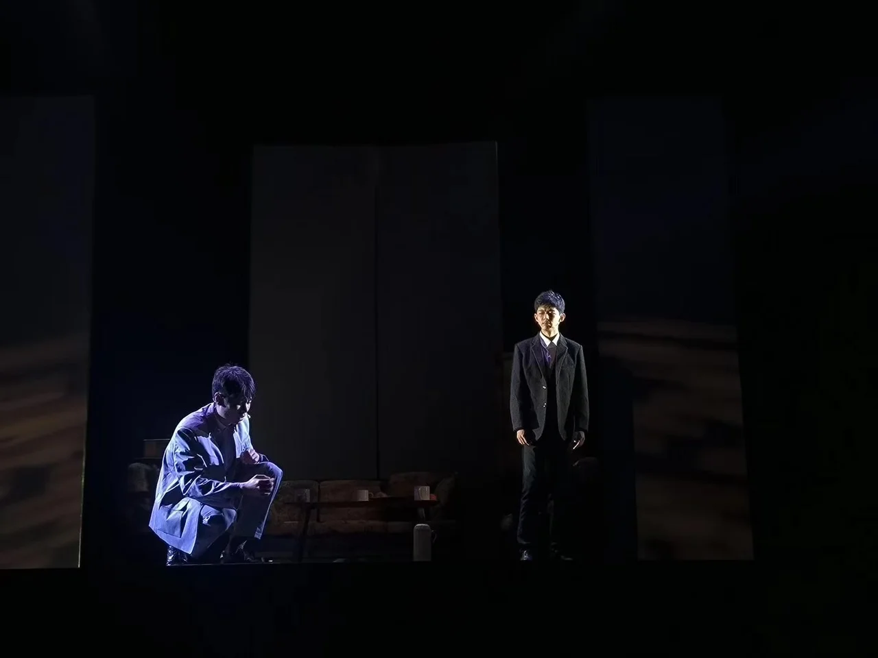On a dark stage, one actor crouches on the left while another in a dark suit stands in the light on the right