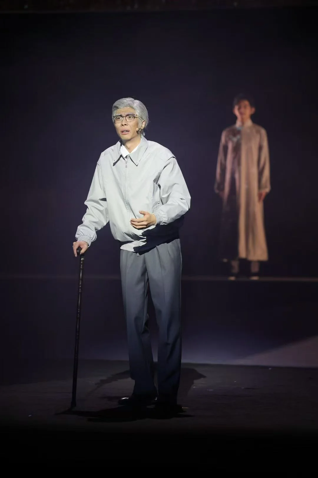 A gray-haired actor wearing glasses and holding a cane, with another performer behind him