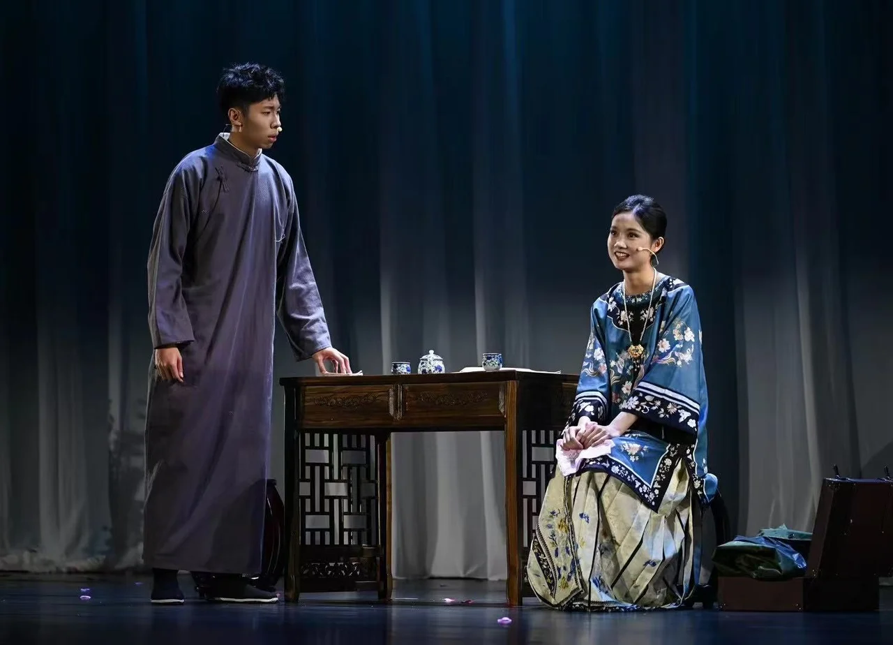An actor in a grey changshan standing to the left of a wooden table, with an actor in blue embroidered clothing seated to the right and a tea set between them
