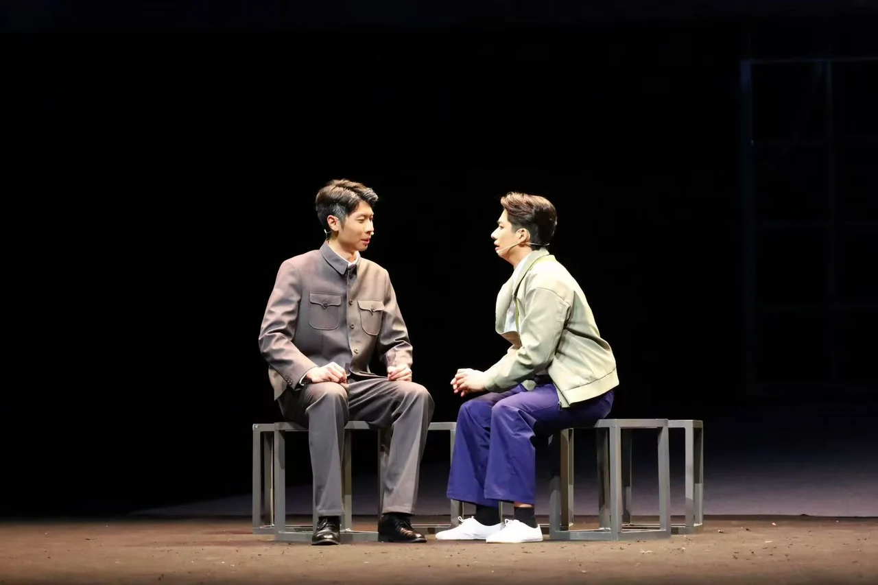 Two actors seated facing one another on square stools on stage