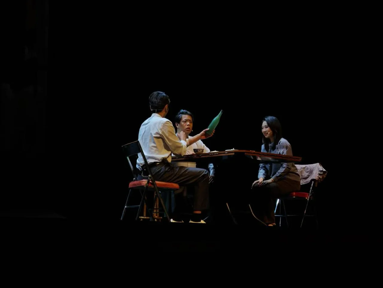 Three actors seated around a table on a dark stage, one holding up a green bottle