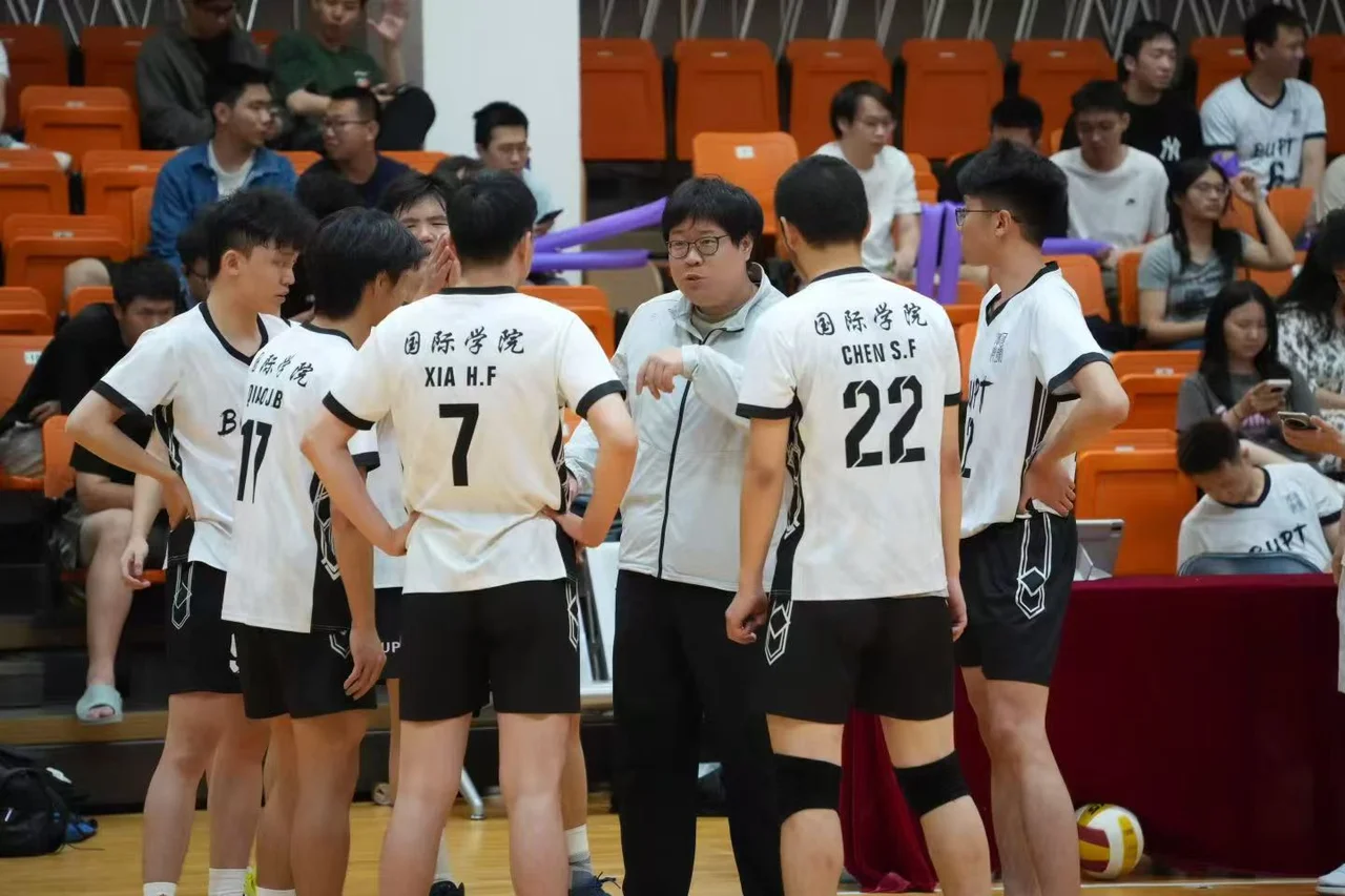 College volleyball players in white International School jerseys gathered around a person in a light jacket beside the court